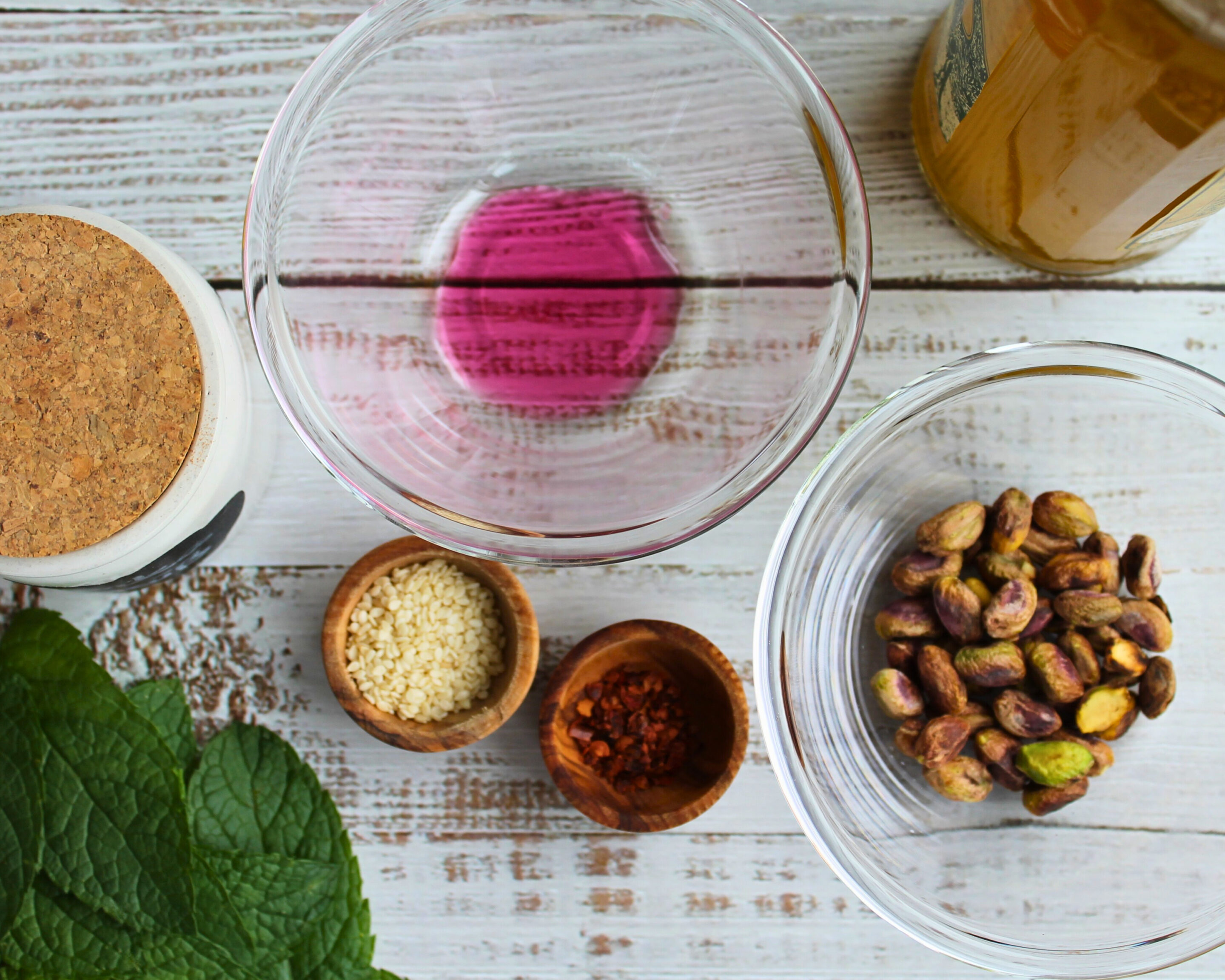zutaten-hummus-schwarze-kichererbsen-pistazien.jpg Übersicht der Hummus-Zutaten: Pistazien, Sesam, Chiliflocken und rosa Zitronensaft in Glas- und Holzschalen.