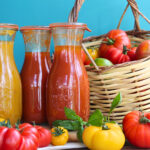 Drei Weckflaschen mit eingekochter Tomatensauce neben einem Korb voller frischer Tomaten und Basilikum.