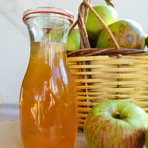 Frisch eingekochter Apfelsaft in einer Glas-Karaffe vor einem Korb mit Äpfeln.