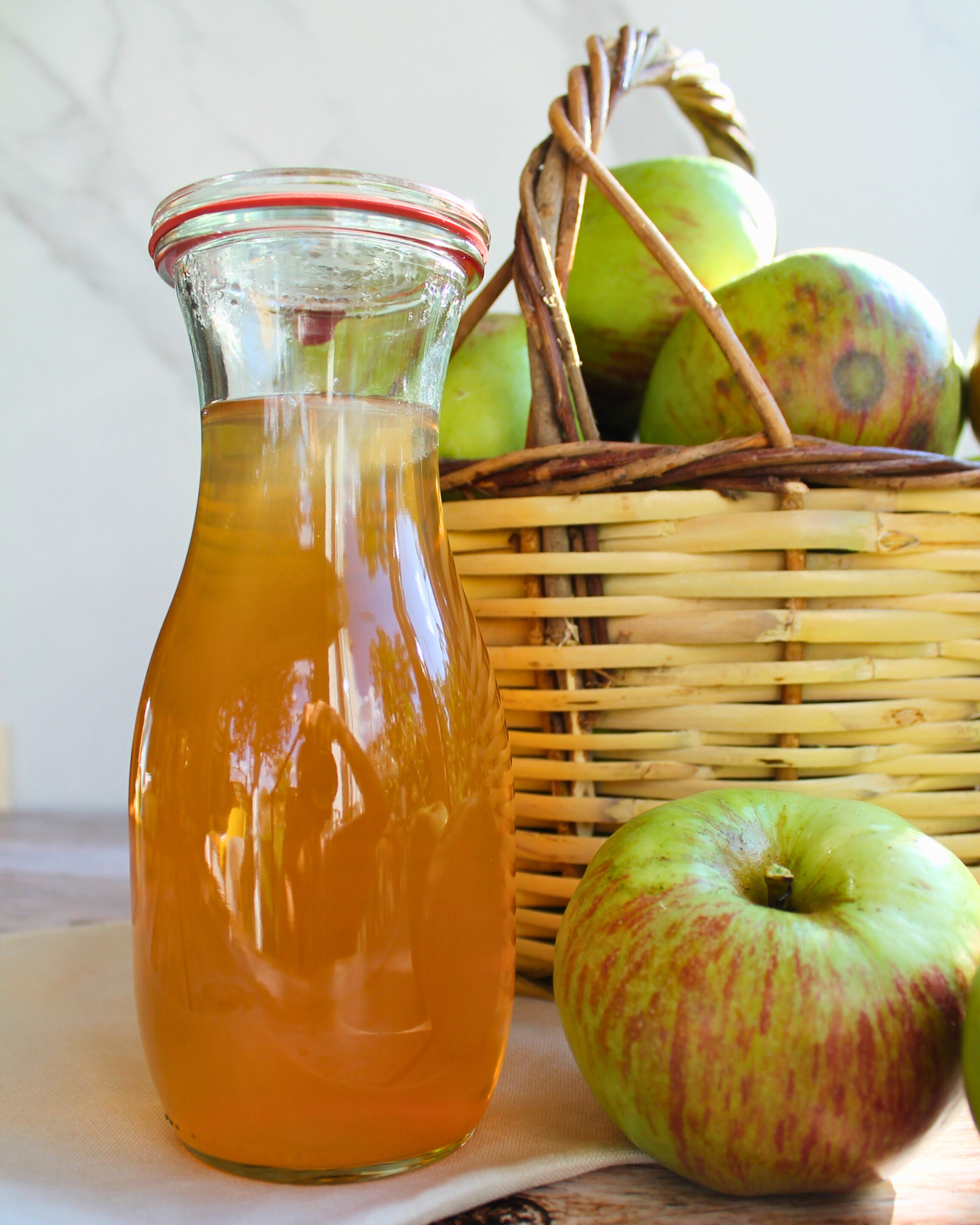 apfelsaft-einkochen-karaffe-aepfel-korb.jpg Frisch eingekochter Apfelsaft in einer Glas-Karaffe vor einem Korb mit Äpfeln.