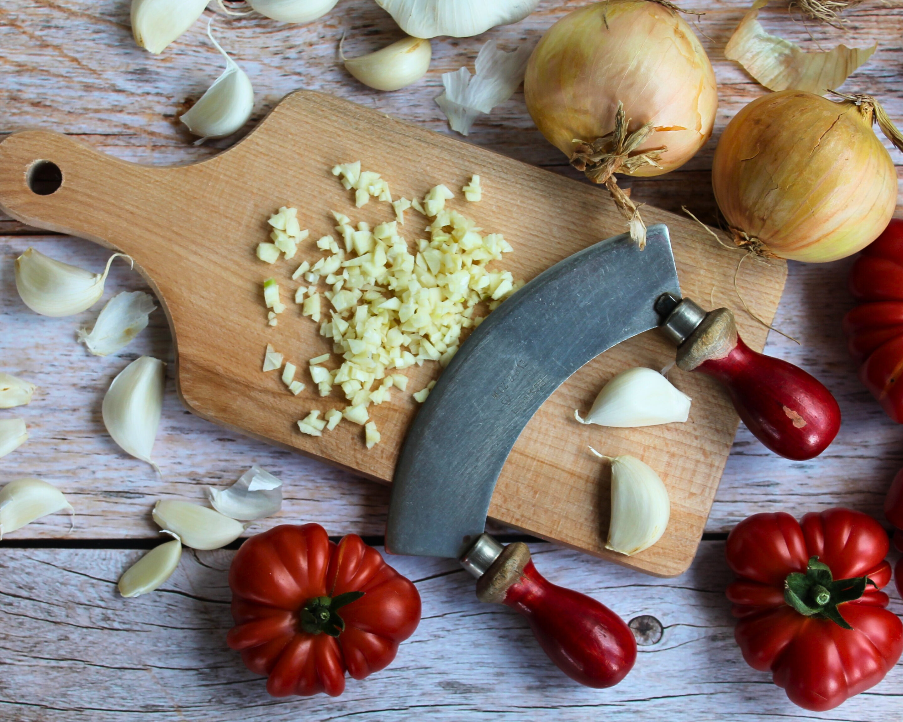 Zwiebeln, Knoblauch und Tomaten auf einem Holzbrett mit Wiegemesser – frische Zutaten für eingekochte Tomatensauce.