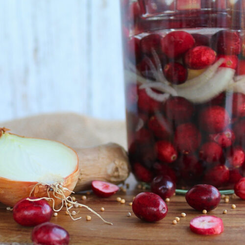 Glas mit fermentierten Cranberries, Ingwer und Zwiebel auf Holzbrett mit verstreuten Senfkörnern.