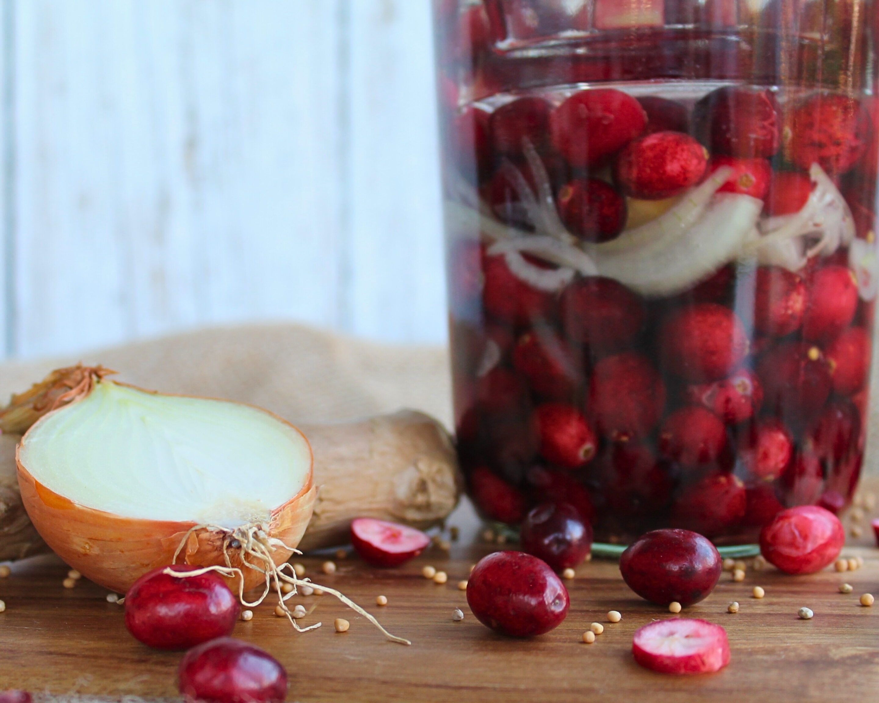 Glas mit fermentierten Cranberrys, Ingwer und Zwiebel auf Holzbrett mit verstreuten Senfkörnern.