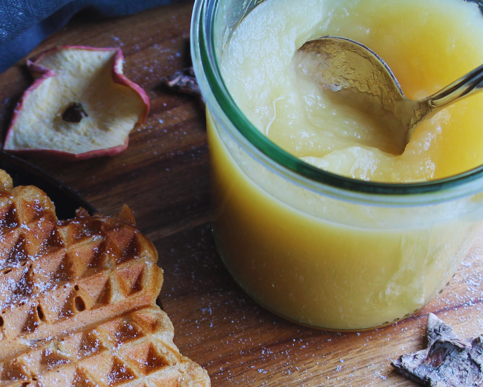 Selbstgemachtes Apfelmus im Glas mit Löffel, daneben Waffeln und getrocknete Apfelscheiben – ideale Geschenkidee aus der Vorratskammer.