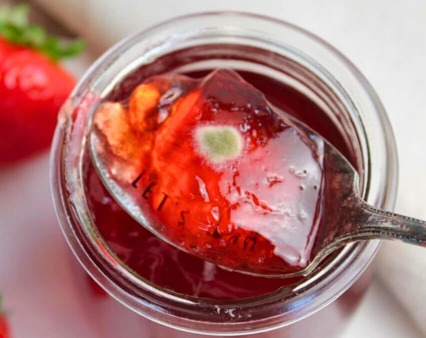 Löffel mit Erdbeermarmelade über geöffnetem Glas, auf der Marmelade ist ein kleiner Schimmelfleck sichtbar.