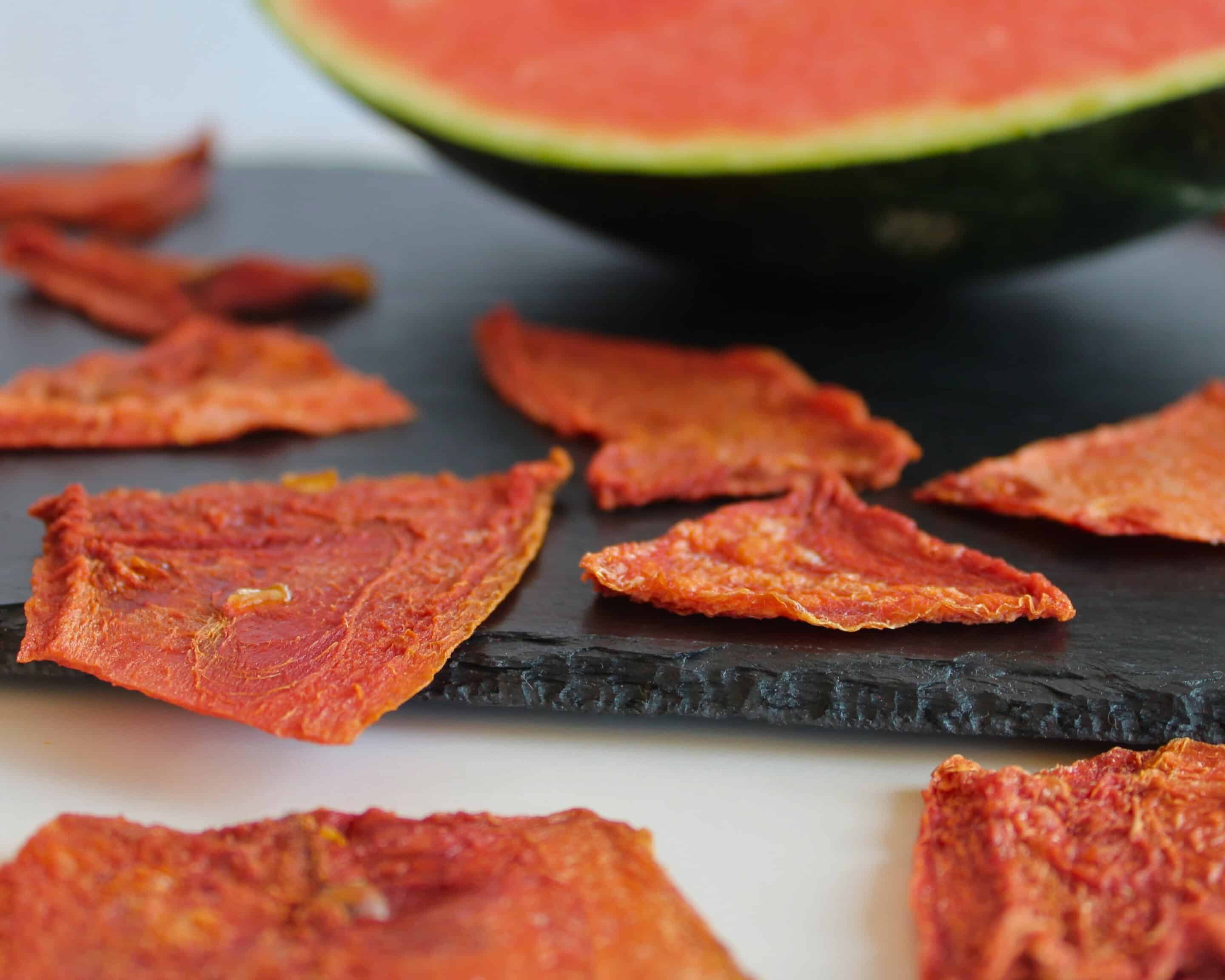Getrocknete Wassermelonen-Chips auf einer Schieferplatte vor einer halbierten Wassermelone